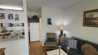This two-floor residence offers a practical and comfortable layout designed for everyday living. The main level showcases warm hardwood flooring that flows throughout the open-concept living and kitchen areas, creating a cohesive and inviting atmosphere.

The living area is furnished with a classic armchair, a striped sofa, and a side table topped with a table lamp, providing a relaxed and functional seating arrangement. A small decorative shelf on the wall adds a subtle personal touch to the space. Natural light filters into the room, keeping the atmosphere bright and airy.

Directly accessible from the living area, the kitchen features dark wood cabinetry extending to the upper walls, a white refrigerator, and efficient use of available space. The open sightline between the kitchen and living room enhances the sense of openness on the main floor.

With its neutral wall tones, hardwood floors, and traditional furnishings, the overall ambiance of the space is warm and understated. The two-floor configuration provides separation between living and private areas, offering a well-organized and functional home environment suitable for a variety of occupants.