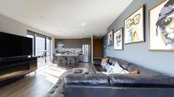 This contemporary single-floor residence showcases a thoughtfully designed open-concept layout that seamlessly blends the living, dining, and kitchen areas into one cohesive space. The living area is anchored by a generous leather sectional sofa, accented with decorative pillows, and complemented by an eclectic gallery wall featuring bold, colorful artwork that adds personality and character to the space.

The kitchen is outfitted with dark, flat-front cabinetry that extends to the ceiling, providing ample storage while maintaining a clean, modern aesthetic. Stainless steel appliances, including a full-size refrigerator and microwave, are integrated seamlessly into the design. A kitchen island with bar seating serves as both a functional prep area and a casual dining spot.

Dark hardwood flooring runs throughout, creating a warm contrast against the light grey walls and white ceiling. Recessed lighting provides even illumination across the space, while expansive floor-to-ceiling windows flood the interior with abundant natural light and offer sweeping views of the surrounding cityscape. The overall ambiance is urban, sophisticated, and modern — a well-appointed space designed for comfortable city living.