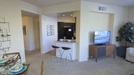 This single-floor space showcases an open-concept layout that integrates the kitchen and living area into a cohesive, functional environment. The kitchen is finished with dark cabinetry that creates a striking contrast against the light-toned walls, and is equipped with black appliances including a refrigerator and built-in microwave. A granite-topped breakfast bar extends from the kitchen, offering casual seating with two sleek, modern bar stools — an ideal spot for dining or entertaining. The bar is styled with stemware and serving bowls, highlighting its practical appeal. The living area features plush carpeting that adds warmth and comfort underfoot, with neutral wall tones contributing to a bright and airy ambiance. A flat-screen television is mounted on a wooden media console, anchoring the living space. A gallery wall of framed artwork adds a decorative touch near the hallway, which leads further into the unit. Recessed lighting and ceiling vents are integrated throughout, ensuring comfort and well-distributed illumination. The overall design balances style and functionality with a clean, contemporary aesthetic.