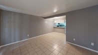 This single-floor space is designed with a practical, open layout that connects the living area to the kitchen, creating a cohesive and functional flow. Light beige ceramic tile flooring runs consistently throughout every room, contributing to a clean and unified aesthetic.

The living area is generously proportioned, featuring neutral gray walls, white baseboards, and a sliding glass door fitted with vertical blinds that allows natural light to filter in. A built-in shelving unit along one wall provides convenient storage and display space. A ceiling-mounted HVAC vent ensures year-round climate control.

The kitchen is positioned adjacent to the living space and is separated by a peninsula counter with a granite-style surface, offering additional prep and casual seating potential. White cabinetry lines the walls above, while the kitchen is equipped with a range, black refrigerator, and an over-the-range microwave. A window above the sink brings in natural light.

The bedroom is a comfortable, quiet room with a window fitted with horizontal blinds and neutral gray walls, consistent with the rest of the unit. The bathroom is bright and finished in white, featuring tile flooring, a bathtub with shower, and standard fixtures, maintaining the clean and functional character of the overall space.
