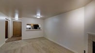 This single-floor residential space offers a clean, functional layout designed for comfortable everyday living. The living area is finished with wall-to-wall carpeting in a neutral tone, creating a warm and inviting atmosphere. Smooth, light-colored walls throughout the space contribute to a bright and airy ambiance, complemented by flush ceiling lighting that evenly illuminates the room. A standout architectural detail is the pass-through window cut into the wall between the living area and the kitchen, fostering an open connection between the two spaces while maintaining a sense of definition. The kitchen, visible through the pass-through, features dark appliances including a refrigerator and range hood, set against light wood cabinetry. A convenient ledge along the pass-through provides additional surface space, ideal for casual dining or serving. The kitchen is also accessible via a separate entryway, offering practical flow between rooms. The overall design is straightforward and well-maintained, with clean lines and neutral finishes that provide a versatile backdrop suitable for a variety of interior styles.