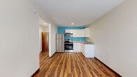 This thoughtfully laid-out single-floor unit makes excellent use of its compact footprint, combining a kitchen and living area into one open, functional space. Rich, warm hardwood-style flooring runs throughout the main area, adding depth and visual continuity. The kitchen is a standout feature, outfitted with stainless steel appliances including a refrigerator, electric range, and over-the-range microwave. White shaker-style cabinetry provides ample storage, while granite-look countertops and a double basin sink add a polished finish. A vibrant teal accent wall behind the kitchen adds a bold pop of color, giving the space a modern, energetic character. Natural light enters through a window in the living area, which also features a wall-mounted HVAC unit for year-round climate control. The entry door, framed with a matching teal border, reinforces the cohesive design aesthetic. The bathroom is clean and contemporary, featuring large-format white floor tiles, a floating vanity with a white countertop and wood-toned base, a framed mirror, and a chrome towel bar. The overall ambiance is fresh, modern, and efficiently organized, making this unit a practical and stylish option.