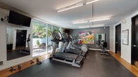 Located at 10306 Meridian Avenue North in Seattle, WA, this single-floor fitness facility offers a thoughtfully designed workout environment that blends functionality with modern aesthetics. The main gym floor is expansive, finished with durable rubber flooring and warm hardwood accents along the perimeter, providing a professional and inviting atmosphere.

The cardio zone is equipped with commercial-grade treadmills, an elliptical, and a stationary bike, all positioned alongside floor-to-ceiling windows that frame views of a serene outdoor pool and lush greenery — flooding the space with natural light. A wall-mounted television and wall clock add practical touches to the workout area. Strength training is well-served by a Precor multi-station cable machine, a dedicated free weights rack with dumbbells, barbells, and weight plates, and a flat bench.

A glass-walled office or training studio sits adjacent to the main floor, outfitted with a desk, seating, and shelving, offering a private space for consultations or programming. Bold pop-art murals add a vibrant, energetic character to the space.

Along the interior wall, a series of black-doored rooms house restrooms, a training salon, and a storage room, keeping the facility organized and fully self-contained. Linear fluorescent lighting throughout ensures consistent illumination across the entire floor.