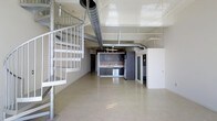 This two-floor loft showcases a striking industrial aesthetic defined by soaring ceilings and prominently exposed silver ductwork that runs throughout the upper portion of the space, adding a raw, urban character. The main level features an open-plan layout with polished concrete-style flooring that reflects light and contributes to a bright, airy atmosphere. A metal spiral staircase on one side of the space serves as both a functional element and a visual focal point, connecting the two floors with architectural flair. The kitchen area is integrated seamlessly into the open floor plan and features dark cabinetry, a stainless steel sink, and standard appliances, offering a functional cooking space without interrupting the flow of the main living area. A partially enclosed room with a glass-panel door is visible toward the rear, providing a private area within the otherwise open layout. The high ceilings amplify the sense of volume and scale throughout the main level, while the clean white walls contrast with the darker kitchen cabinetry and metallic industrial details. Overall, the space balances urban industrial design with practical modern finishes.