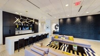 This contemporary amenity space is designed to impress, offering a seamlessly connected series of social and recreational areas across a single floor. The entrance greets visitors through sleek black-framed glass doors that open into a bright, tile-floored lobby anchored by a dramatic textured black accent wall and lush indoor greenery. A glass-enclosed office or leasing suite adds transparency and visual depth to the entry corridor.

The central lounge is a standout, featuring soaring ceilings clad in large-format tile and concrete-toned finishes, with a bold abstract artwork as the focal point. An eye-catching arrangement of sculptural pendant lighting — including geometric and branching brass fixtures — illuminates plush gray sofas accented with gold and yellow throw pillows, set atop a graphic black-and-white striped rug. Adjacent to the lounge is a sleek bar counter with dark cabinetry, a bright white surface, and bar seating beneath a striking multi-arm chandelier.

The game room offers a more relaxed atmosphere, equipped with a full-size billiards table and a shuffleboard table, set on patterned carpet with natural light streaming through large windows. A separate lounge nook within the game room area includes a linear fireplace, modern dark leather seating, and additional accent walls in deep navy blue, creating a cozy retreat. Throughout the space, curated artwork, potted plants, and contemporary furnishings contribute to a polished, upscale ambiance.