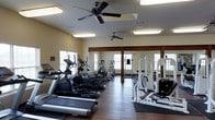This fitness center offers a comprehensive workout environment designed to accommodate a wide range of exercise needs. The space features rich hardwood flooring throughout, providing a durable and attractive surface well-suited for both cardio and strength training activities. Multiple ceiling fans are mounted overhead, ensuring consistent air circulation during workouts, while recessed and flush-mounted lighting keeps the space bright and well-lit at all times. Large windows along the walls invite generous amounts of natural light, contributing to an open and energizing atmosphere. A full wall of mirrors reflects the space, making the room feel expansive while also allowing users to monitor their form during exercise. The equipment lineup includes a row of treadmills, multiple elliptical cross-trainers, and a variety of strength and resistance machines, offering a thorough selection for both cardiovascular and muscle-building workouts. Sturdy wooden support columns add a touch of rustic character to the otherwise modern facility. The overall layout is thoughtfully organized to maximize floor space and allow comfortable movement between stations, making this fitness center a practical and inviting space for daily exercise routines.