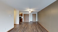 This single-floor residence offers a functional and well-organized layout designed for comfortable everyday living. The main living area is finished with rich, dark hardwood-style flooring that extends throughout the space, lending warmth and a cohesive aesthetic. A ceiling fan with an integrated light fixture is centrally positioned overhead, providing both air circulation and ambient lighting. The room features a neutral color palette, with crisp white walls complemented by a warm taupe accent wall, creating a subtle visual contrast that adds depth to the space. A set of sliding wood-panel closet doors provides convenient built-in storage, while a solid wood entry door with a modern finish serves as the welcoming focal point of the main wall. The open layout allows for a clear sightline into the adjacent kitchen, which is equipped with dark wood cabinetry, a black range, and an overhead ventilation hood. The kitchen's compact and efficient design makes it easily accessible from the living area. The overall ambiance is clean, neutral, and move-in ready, offering a practical foundation for personalization and furnishing.