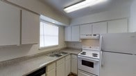 This single-floor space includes a well-organized kitchen designed for everyday functionality. The kitchen features white cabinetry that extends to the upper walls, providing generous storage space. A double-basin sink is positioned beneath a window fitted with horizontal blinds, allowing natural light to filter into the space. The countertops are finished in a speckled laminate surface, offering practical workspace along the perimeter of the kitchen in an L-shaped layout. White appliances include a coil-burner electric range with an overhead ventilation hood, and a full-sized refrigerator positioned in the corner. A dishwasher is partially visible at the base of the cabinetry. Overhead, a fluorescent light fixture provides bright, even illumination across the workspace. The lower cabinetry offers additional storage with a consistent white finish that matches the upper cabinets, creating a cohesive and clean aesthetic throughout. The overall design is straightforward and utilitarian, prioritizing practicality and ease of use within a compact, efficiently arranged layout.