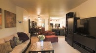 This single-floor space is designed around an open-concept floor plan that seamlessly connects the living room, dining area, and kitchen into one cohesive, flowing environment. The living room anchors the space with plush seating, including a deep burgundy sectional sofa with an ottoman and a neutral-toned couch, arranged around a decorative mosaic-top coffee table adorned with a fresh floral centerpiece. Warm ambient lighting from a ceiling fan fixture overhead lends the space a cozy, inviting atmosphere. A large black shelving unit along the right wall provides substantial storage and display space. Moving through the open corridor, the dining area is visible to the left, featuring a wooden dining table surrounded by chairs beneath soft overhead lighting. The kitchen sits at the rear of the space and is equipped with visible countertops, cabinetry in contrasting light and dark tones, and modern appliances including a microwave. A skylight or recessed ceiling light brightens the kitchen area. The consistent use of warm tones, carpet flooring, and open sightlines throughout the space contribute to a relaxed and functional living environment.