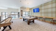 This contemporary game room and amenity lounge is thoughtfully designed for social recreation and casual gatherings. The space is anchored by a full-size pool table positioned centrally beneath an elegant drum pendant light with gold accents, providing warm, focused illumination over the playing surface. A shuffleboard table complements the gaming setup, offering additional entertainment options.

One wall serves as a standout design feature, finished in a soft sage green and adorned with a decorative rope grid pattern that adds visual texture and an artistic focal point. A wall-mounted flat-screen TV is integrated into this feature wall, flanked by a built-in wooden counter with modern bar-height seating — ideal for spectators or casual socializing.

Underfoot, a patterned loop carpet in neutral tones adds comfort and visual interest throughout. Recessed ceiling lighting supplements the pendant fixture, ensuring the room is well-lit. Large windows along one side of the room invite natural light and provide views of the surrounding exterior, enhancing the open, airy atmosphere.

A cable-railing mezzanine overlook and adjacent corridor connect this space to the broader amenity floor, which features clean white walls, warm wood accents, and glass-paneled double doors — reinforcing the modern, upscale character of the overall facility.