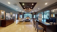 This single-floor community clubhouse or leasing center is thoughtfully designed to blend sophistication with functionality. The main lounge area serves as the centerpiece, featuring polished cream tile flooring, deep charcoal-gray walls with crisp crown molding, and a striking coffered ceiling with a dark inset panel anchored by a multi-arm chandelier. Recessed lighting complements the natural light streaming through large arched and French-style windows, which frame views of lush greenery outside.

The lounge is furnished with cream sofas, upholstered accent chairs, and a dark wood coffee table set atop layered area rugs, creating comfortable seating arrangements for both social gatherings and quiet relaxation. A round dining table with modern chairs occupies a secondary seating nook, while a dedicated reception or leasing desk area features nailhead-trimmed chairs suited for consultations.

Adjacent to the lounge, a fitness center is visible through glass partitions, outfitted with cardio equipment and mirrored walls. A kitchenette or beverage station with warm wood cabinetry adds convenience. Decorative wall art and curated accessories throughout contribute to the polished, welcoming ambiance of the overall space.