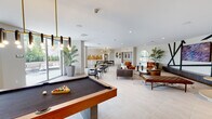 Located at 1110 W Flagler St in Miami, FL, this single-level amenity space offers a well-appointed communal environment designed for both relaxation and social interaction. The open-plan layout is unified by large-format light tile flooring and crisp white ceilings with recessed lighting, creating a bright and airy atmosphere throughout.

The space is divided into several distinct functional zones. A game area centers around a full-size billiards table, flanked by tufted leather benches and teal upholstered booth seating. Adjacent to this is a lounge area featuring a bold accent wall with angular dark steel geometric detailing framing a large wall-mounted display, accompanied by cognac leather chairs, a plush sofa, and layered area rugs that add warmth and texture.

A dedicated dining zone includes a dark communal table with modern chairs, positioned beneath a sculptural brass and black chandelier. The nearby kitchenette features white cabinetry, a stainless steel refrigerator, and a distinctive wood-tile geometric backsplash that adds an artistic focal point. Arched windows and expansive glass doors introduce generous natural light and views of lush greenery, enhancing the overall inviting ambiance of the space.