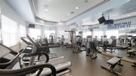 This fitness center is a thoughtfully designed space built to support a comprehensive workout experience. The layout is open and spacious, with a tray ceiling and recessed lighting that creates a bright, energizing atmosphere. Large windows along one wall invite natural light, enhancing the overall sense of openness throughout the room.

The facility is outfitted with a robust selection of Life Fitness cardio equipment, including treadmills positioned toward the front of the space for easy access. A variety of strength training machines are arranged throughout the room, offering options for both upper and lower body workouts. The flooring features durable, dark rubberized material suited for high-traffic athletic use.

Floor-to-ceiling mirrors line one wall, making the space feel even larger while allowing users to monitor their form during exercise. A wall-mounted television is visible, providing entertainment during workouts. The motivational word "PREVAIL" is prominently displayed on the mirrored wall, setting an inspiring tone for the space.

The ceiling design incorporates a raised tray detail with crown molding, lending a polished, upscale finish that elevates the aesthetic above a standard gym environment. Overall, this fitness center balances functionality with a clean, modern design to create an inviting and well-appointed workout destination.