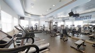 This fitness center is a thoughtfully designed space built to support a comprehensive workout experience. The layout is open and spacious, with a tray ceiling and recessed lighting that creates a bright, energizing atmosphere. Large windows along one wall invite natural light, enhancing the overall sense of openness throughout the room.

The facility is outfitted with a robust selection of Life Fitness cardio equipment, including treadmills positioned toward the front of the space for easy access. A variety of strength training machines are arranged throughout the room, offering options for both upper and lower body workouts. The flooring features durable, dark rubberized material suited for high-traffic athletic use.

Floor-to-ceiling mirrors line one wall, making the space feel even larger while allowing users to monitor their form during exercise. A wall-mounted television is visible, providing entertainment during workouts. The motivational word "PREVAIL" is prominently displayed on the mirrored wall, setting an inspiring tone for the space.

The ceiling design incorporates a raised tray detail with crown molding, lending a polished, upscale finish that elevates the aesthetic above a standard gym environment. Overall, this fitness center balances functionality with a clean, modern design to create an inviting and well-appointed workout destination.