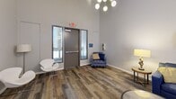 This 720 square foot single-floor space in Los Alamitos, CA, is a versatile and contemporary communal area designed for multiple uses. The interior boasts high ceilings with modern globe pendant lighting, creating an open and airy ambiance. The space is divided into distinct functional areas, including a lounge, game room, and meeting space.

The lounge area features a comfortable navy blue sectional sofa accented with yellow and patterned throw pillows, complemented by a sleek glass coffee table and artistic wall decor. Adjacent to this, a game area includes a foosball table, adding a recreational element to the space.

A meeting area is equipped with a round table and modern chairs, ideal for small gatherings or work sessions. Large windows allow ample natural light and offer views of the surroundings. The room also includes a wall-mounted TV and additional seating options, such as modern armchairs in various styles.

The flooring throughout is a durable wood-look material in warm, varied tones, unifying the different areas while adding a touch of warmth. The walls are painted in a neutral light gray, serving as a backdrop for colorful abstract artworks that add visual interest and personality to the space.

Practical features include an exit door with safety signage and what appears to be a small kitchenette or service area. The overall design strikes a balance between functionality and style, creating a welcoming environment for various activities and gatherings.