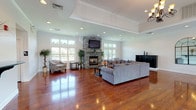 This generously proportioned single-floor space is designed for both relaxation and social gatherings, offering a variety of functional zones within an open, flowing layout. Rich, high-gloss hardwood flooring runs throughout, reflecting the abundant natural light that streams in through large windows lining the walls.

The main living area serves as the heart of the space, anchored by a dramatic floor-to-ceiling stacked stone fireplace with a mounted flat-screen television above. Comfortable sectional sofas and accent chairs are arranged to create an inviting lounge environment. Elevated tray ceilings with recessed lighting add architectural depth and an airy sense of height to the room.

Adjacent to the lounge, the space opens into a recreation and dining zone, complete with a billiards table and a formal dining setup featuring dark wood furniture. A chandelier overhead lends a touch of elegance to this area. French doors lead to an outdoor view, reinforcing the bright, open ambiance throughout.

The kitchenette offers warm wood cabinetry, dark granite countertops with an undermount sink, and tile flooring — providing practical amenities to support events and gatherings. Throughout the space, a neutral color palette, crown molding, and thoughtful architectural details contribute to a polished yet welcoming atmosphere.