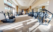 This 2,050-square-foot fitness center, located in the Marketplace community of Irvine, CA 92602, is a single-floor facility thoughtfully designed to support a comprehensive workout experience. The space features an open, rectangular layout with high ceilings, recessed linear lighting, and a neutral color palette of warm beige and cream tones accented by natural wood slatted columns that add architectural warmth throughout.

Floor-to-ceiling windows and large glass doors flood the interior with abundant natural light and provide expansive views of the surrounding palm tree-lined landscape, creating an energizing yet serene atmosphere. The patterned carpet tile flooring defines the training zones while contributing to noise reduction across the space.

The cardio zone is well-equipped with treadmills, stationary bikes, elliptical trainers, stair climbers, step mills, and rowing machines. The strength training area features a cable machine, multiple selectorized strength machines, a full dumbbell rack with benches, and dedicated free weight stations. Large mirrors along the walls enhance spatial perception and support form monitoring during workouts.

A vibrant abstract mural in pastel tones serves as a striking focal point on one wall, accompanied by a mounted flat-screen TV and a ceiling fan, adding comfort and visual interest. The overall design balances functionality and modern aesthetics, making it a well-appointed fitness amenity.