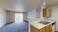 Located at 8100 N Thomas Meyers Dr in Kansas City, MO, this single-story unit offers a practical, open layout designed for comfortable everyday living. The living area is spacious and carpeted in blue-gray tones, with a large sliding glass door fitted with vertical blinds that allows natural light to fill the room while providing access to an outdoor balcony area. A textured ceiling and a wall-mounted heating unit add functional character to the space.

The kitchen connects directly to the living area via a raised breakfast bar with a built-in sink, creating an open, semi-divided layout. Warm oak-toned cabinetry lines the kitchen walls, offering generous storage above and below the countertops. White appliances — including a coil-top electric range, refrigerator, and dishwasher — are paired with light laminate countertops. The kitchen floor features wood-look laminate flooring, which also carries into the bathroom.

The bedroom is a quiet, carpeted room with a window overlooking outdoor greenery, providing a comfortable and naturally lit retreat. The bathroom includes a full tub and shower combination with white tile surround, along with standard fixtures. Overall, the unit presents a clean, functional layout well-suited for everyday living.