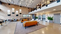 Located at 207 S Maple St in Mount Prospect, Illinois, this contemporary lobby is a striking welcome space defined by its dramatic double-height ceilings and open, airy layout. The design masterfully blends industrial and modern aesthetics, anchored by a prominent exposed brick feature wall adorned with curated artwork. Polished concrete floors reflect the abundant ambient light cast by a sculptural geometric pendant light installation overhead, creating a dynamic visual centerpiece.

The main seating area is furnished with bold cognac leather sofas, patterned accent ottomans, and neutral lounge chairs arranged on a herringbone-patterned area rug, offering a comfortable and stylish gathering space. An upper-level mezzanine with sleek metal railings overlooks the lobby, adding architectural depth and a sense of vertical scale.

On one side, floor-to-ceiling gridded windows flood a casual café-style nook with natural light, providing street views and a relaxed seating area with banquette and bistro furnishings. Adjacent to this, a pass-through window with bar seating opens to a neighboring retail or food service space. The elevator bank is neatly integrated along a wood-paneled wall, flanked by lush tropical planters that add warmth and life to the polished surroundings.