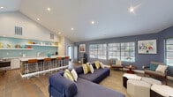 Located at 2645 Stone Creek Dr in Sacramento, California, this single-floor community lounge seamlessly combines a fully equipped kitchen with a welcoming living area in one open, well-designed layout. The space is defined by its soaring vaulted ceilings, which lend an airy, expansive feel throughout.

The kitchen area is a standout feature, anchored by a large island with a crisp white quartz countertop and reclaimed wood paneling, providing ample seating with modern bar stools. The counters along the back wall are outfitted with stainless steel appliances, including a dishwasher and oven, complemented by white shaker cabinetry. A vibrant light-blue accent wall with open floating shelves displaying colorful décor adds a playful, inviting character to the space.

The living area transitions naturally from the kitchen, centered around a large sectional sofa in a deep navy hue, accessorized with yellow and green accent pillows. A large wall-mounted flat-screen TV serves as the focal point on a textured feature wall, while modular ottoman seating offers flexible arrangement. Multiple windows with plantation shutters allow ample natural light and contribute to the bright, welcoming ambiance. Warm hardwood flooring runs throughout, unifying the space. A glass door provides a view into an adjacent fitness area, adding further functionality to this communal retreat.