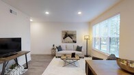 This single-floor, one-bedroom, one-bathroom residence offers a thoughtfully designed layout that maximizes its well-proportioned footprint. The living area serves as the centerpiece of the home, featuring smooth white walls, recessed lighting, and wood-look flooring that runs throughout, creating a cohesive and polished look. A large window fitted with horizontal blinds allows natural light to fill the room, enhancing the bright and airy atmosphere. The clean-lined aesthetic is complemented by neutral tones, giving the space a contemporary yet welcoming feel. The open layout provides flexibility for furniture arrangement, as demonstrated by the comfortable seating area complete with a light-toned sofa, decorative accent pillows, a textured area rug, and an industrial-style coffee table. Tasteful wall art adds a personal touch without overwhelming the minimalist design. Recessed ceiling fixtures ensure consistent, even illumination throughout the day and evening. Overall, the space presents a modern, well-maintained environment with functional design elements suited for comfortable everyday living.
