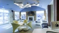 This single-floor space showcases a bold and imaginative contemporary interior design that immediately commands attention. The vaulted ceiling is painted a vivid periwinkle blue and serves as a dramatic backdrop for a sculptural, wave-shaped pendant light fixture that stretches across the room, casting a bright, ambient glow throughout the space. Large windows on multiple walls flood the interior with abundant natural light, offering views of the surrounding outdoor environment and enhancing the open, airy atmosphere. At the heart of the room stands a classic white fireplace, crowned by a striking fan-shaped metallic wall sculpture that adds an artistic focal point. The furnishings are distinctly futuristic and avant-garde — curvaceous, high-gloss chartreuse pod chairs and smooth, rounded white ottomans are paired with patterned upholstered seating and sleek side tables, creating a dynamic yet cohesive aesthetic. Warm tan walls contrast beautifully with the cool blue ceiling, while the light-toned tiled floor ties the composition together. The overall ambiance is one of creative sophistication — a thoughtfully designed space that balances artistic expression with functional lounge comfort.