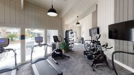 The fitness center at Dwell at Greenridge, located in North Charleston, South Carolina, is a well-appointed, single-floor facility designed to support a comprehensive workout experience. The space is defined by its striking architectural character, featuring soaring vaulted ceilings with exposed beam detailing and white shiplap wall paneling that lend a clean, modern-farmhouse aesthetic throughout.

Large floor-to-ceiling windows and glass entry doors flood the interior with abundant natural light, offering views of the surrounding community and creating an open, airy atmosphere. The flooring transitions between durable carpet tile in the cardio and strength zones, providing both comfort and practicality underfoot.

The cardio area is well-equipped with treadmills, ellipticals, and stationary bikes — including a Peloton-style cycle — all positioned for optimal use of the space. Wall-mounted flat-screen televisions are positioned throughout for entertainment during workouts. A dedicated strength area features a multi-use cable machine, a full dumbbell rack, and additional free weights to accommodate a range of training needs.

Pendant lighting fixtures hang from the vaulted ceiling, complementing the natural light and adding to the inviting ambiance. A hydration station and sanitation supplies are conveniently available near the exit, which opens directly to the outdoor community grounds. The "Dwell at Greenridge" branding is prominently displayed on the wall, reinforcing the community identity throughout the space.