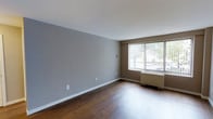 This single-floor space offers a clean, functional layout with a straightforward design aesthetic. The main living area is finished with rich, dark hardwood flooring that provides a warm contrast to the cool grey and taupe-toned walls. White baseboards run along the perimeter, adding a crisp, defined border between the walls and floor.

A large window dressed with vertical blinds serves as a focal point along one wall, allowing a generous amount of natural light to enter the room while offering views of the surrounding outdoor environment. A baseboard heating unit is positioned beneath the window, providing an efficient heating source integrated into the room's layout.

The walls are painted in a neutral grey palette, creating a versatile backdrop suitable for a variety of décor styles. The space feels open and uncluttered, with a simple, practical design that emphasizes functionality. Electrical outlets are conveniently placed along the walls, and the overall condition of the space appears well-maintained. The combination of natural light, hardwood flooring, and neutral tones gives the room a balanced and livable ambiance.
