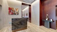 This refined building lobby presents a seamless blend of contemporary design and upscale materials across a generously proportioned single-floor layout. The entrance greets visitors through floor-to-ceiling glass doors framed in brushed steel, opening onto travertine-tiled floors that extend throughout the space. A striking dark marble reception desk with brass detailing anchors the entry area, positioned alongside curated, colorful artwork that animates the neutral walls.

The corridors are lined with warm, richly grained wood wall panels and recessed cove lighting, creating an inviting transition toward the elevator banks. Two distinct lounge areas offer comfortable seating arrangements: one features bold red velvet armchairs clustered around a round marble coffee table, while the other showcases a tufted charcoal sofa paired with cream accent chairs and a geometric glass-top table. Both lounges are bathed in abundant natural light from expansive floor-to-ceiling windows dressed with sheer draping curtains, offering views of surrounding greenery and the urban streetscape beyond.

A decorative fireplace with a dark marble surround and chevron-patterned screen adds architectural interest to one lounge. Throughout the space, original artwork, sculptural accessories, and carefully selected furnishings contribute to an atmosphere of understated elegance befitting a premier professional or residential building.