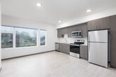 This single-floor residential unit offers a clean, contemporary design characterized by light wood-look flooring, neutral wall tones, and consistent wood-grain cabinetry throughout. The kitchen is thoughtfully outfitted with full-height cabinetry in a warm taupe wood-grain finish, stainless steel appliances including a range, over-the-range microwave, and refrigerator, along with a white countertop and undermount sink — providing both functionality and a modern aesthetic. An adjacent nook with additional cabinetry and a second refrigerator offers flexible storage and utility options. The bedroom features plush wall-to-wall carpeting and a wide window that invites natural light and views of the surrounding neighborhood. The bathroom is efficiently designed, featuring a single-sink vanity with matching wood-grain cabinetry, a full soaking tub with shower, and a conveniently integrated stacked washer and dryer unit — eliminating the need for shared laundry facilities. The hallway connecting the rooms is well-lit with recessed lighting and lined with wood-tone doors, culminating in a bright window at the end that draws in additional natural light. The overall layout, as illustrated by the floor plan, is compact yet functional, with a logical flow between living spaces.