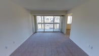 This single-floor residence centers around a spacious, open-concept living area finished with plush wall-to-wall carpeting and neutral-toned walls that create a clean, versatile backdrop. The most prominent feature of the main living space is a large sliding glass door system flanked by floor-to-ceiling curtain panels, which floods the room with natural light and provides direct access to a private balcony. From the balcony, views of the adjacent building's pitched roofline and surrounding trees add a sense of depth and connection to the outdoors. A secondary window positioned to the right of the main living area further enhances the natural light throughout the space. The open layout allows for flexible furniture arrangements and a comfortable sense of spaciousness. An adjoining room, visible through an open doorway to the right, extends the functional living area and appears to benefit from its own natural light source. White baseboards and a smooth ceiling complete the interior, lending a simple, understated finish. Overall, the space offers a bright, airy atmosphere with practical indoor-outdoor connectivity.