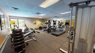 Located at 801 Cooper St in Deptford Township, NJ, this well-equipped fitness center offers a comprehensive workout environment on a single floor. The space features durable rubber-tile flooring throughout, providing both safety and comfort during exercise. A drop ceiling with recessed fluorescent lighting ensures consistent, bright illumination across the entire area.

The cardio section is impressively stocked with multiple treadmills, elliptical trainers, and a recumbent bicycle, catering to a variety of aerobic workout preferences. Large wall-to-wall mirrors line one side of the room, enhancing the sense of space and allowing users to monitor their form. The strength training area includes a cable machine, a dumbbell rack with a full range of weights, and adjustable workout benches, supporting a thorough resistance training routine.

Natural light filters in through several windows, and a sliding glass door opens to a view of the outdoor pool area, creating a bright and motivating atmosphere. Accent walls in warm yellow add a lively touch to the otherwise neutral color palette of gray and white. The space is thoughtfully arranged to maximize floor space and equipment accessibility, making it a functional and welcoming facility for users of all fitness levels.