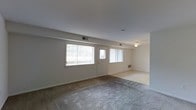 This single-floor residence offers a straightforward and functional layout designed for comfortable everyday living. The main living area is generously sized, featuring wall-to-wall carpet that adds warmth underfoot. Large windows along the exterior wall flood the space with natural light, while a door provides direct access to the outdoors. A ceiling fan is visible in the adjoining area, which flows seamlessly from the main room, creating an open and connected feel. Neutral wall tones throughout lend the space a clean, understated appearance.

The kitchen is efficiently designed, making excellent use of its footprint with an L-shaped layout. Rich, dark wood cabinetry extends from counter height to the ceiling, providing ample storage space. Light-colored countertops offer a contrasting brightness to the cabinetry. The kitchen is equipped with a coil-burner electric range with an overhead exhaust hood, a stainless steel sink, a dishwasher, and a full-size refrigerator. Overhead lighting ensures the workspace is well illuminated. Together, the living and kitchen areas present a practical, move-in-ready space suited for a range of lifestyles.
