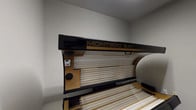 This dedicated tanning room is a compact, self-contained space designed specifically for indoor tanning sessions. The centerpiece is a Montego Bay tanning bed, a full-sized unit featuring dual UV lamp arrays on both the upper canopy and lower base, finished in a sleek black and gold design. The bed is positioned to make efficient use of the room's available floor space.

The room is finished with neutral gray walls and carpeted flooring, creating a clean and functional environment. A private entry door features a chain lock for added security during sessions, along with a wall-mounted hook for hanging clothing or personal items. Adjacent to the door, a wall-mounted paper towel or sanitation product dispenser is conveniently accessible for hygiene purposes.

A full-length mirror is positioned along one wall, allowing users to check their appearance before and after sessions. An informational chart is mounted on the wall, likely providing session guidelines or tanning schedules for user reference. The overall layout is straightforward and efficiently organized, prioritizing practicality and privacy. The room's simple design keeps the focus on the tanning equipment while maintaining a clean, professional atmosphere suitable for a salon or fitness facility setting.