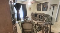 This single-floor space showcases a thoughtfully designed living area that exudes classic elegance and refined taste. The living room serves as the centerpiece, featuring ornate wooden furniture with intricate carved detailing, including a cushioned sofa adorned with decorative pillows in rich earth tones and a matching set of armchairs that invite comfortable seating. A round wooden coffee table anchors the seating arrangement, resting atop a bold patterned area rug that adds depth and contrast to the light-toned tile flooring beneath. Floor-to-ceiling dark drapery panels frame the windows, allowing for both dramatic styling and effective light control, while natural light filters in to brighten the space when desired. A ceiling fan with an integrated light fixture provides both function and a finishing architectural touch overhead. The walls are adorned with a curated collection of framed artwork, lending a gallery-like quality to the room. A dark wood display cabinet or armoire along the wall adds both storage and visual weight to the overall composition. The space projects a warm, sophisticated atmosphere with its layered textures, classic furnishings, and carefully selected décor elements.