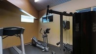 This single-floor fitness room offers a well-equipped exercise space designed for varied workout routines. The room features a vaulted, wood-paneled ceiling that adds architectural character and contributes to an open, airy feel. Warm amber-toned walls provide a energizing atmosphere, while a dark rubberized floor covering offers both durability and safety underfoot. Natural light enters through two horizontal windows positioned along the walls, keeping the space bright without compromising privacy. A full multi-station cable machine anchors one side of the room, complete with a pulley system and weight stack, making it suitable for a wide range of strength training exercises. An elliptical cross-trainer provides low-impact cardiovascular options, and additional free weights and equipment are visible throughout the space. Wall-mounted mirrors along one side visually expand the room and allow users to monitor their form during exercise. The compact yet functional layout makes efficient use of the available space, providing a comprehensive home fitness environment suitable for both strength and cardio training.