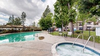 The outdoor pool area at 6736-6756 N Clybourn Avenue in North Hollywood, Los Angeles, serves as a central communal amenity within the residential community. The generously sized rectangular pool features clear, turquoise water and is complemented by a wide concrete deck that provides ample space for lounging and movement around the perimeter.

The area is enclosed by a black wrought iron fence, offering both security and an open, airy feel that maintains sightlines to the surrounding landscape. Mature trees, including tall pines and broad-canopied deciduous varieties, frame the space on multiple sides, providing natural shade and a sense of seclusion while softening the built environment. Lush shrubs and greenery line portions of the fencing, adding additional privacy and visual appeal.

Pool access points include entry steps with metal handrails, and safety equipment such as a life ring is visibly mounted near the pool's edge. Seating is available on the surrounding deck, and informational signage is posted at the entry gate. The broader community buildings are visible in the background, integrating the pool area naturally within the residential setting. The overall atmosphere is relaxed and well-maintained, ideal for outdoor recreation in the Southern California climate.