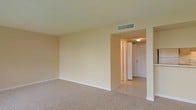 This single-story space presents a clean, functional layout with a neutral color palette throughout. The main living area features wall-to-wall carpet in a warm beige tone, complemented by matching cream-colored walls that lend a cohesive, understated aesthetic to the interior. A textured ceiling adds subtle visual interest overhead, while a central HVAC vent ensures efficient climate control throughout the space.

An open doorway connects the carpeted living area to an adjacent tiled section, which appears to serve as an entryway or transitional zone. This area features light-toned ceramic tile flooring, providing a practical and durable surface near what appears to be a front entry door. A flush-mount ceiling light fixture illuminates this zone, and a bifold or panel door is visible along the wall, likely providing access to a closet for additional storage.

White baseboards run along the perimeter of both areas, offering a crisp contrast to the neutral walls and flooring. Electrical outlets are positioned conveniently along the walls. The overall ambiance is bright, clean, and move-in ready, offering a blank canvas well-suited for a variety of furnishing and decorating styles.