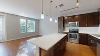 This single-floor residence offers a thoughtfully designed open-concept layout that seamlessly connects the kitchen and living areas. The kitchen is a standout feature, showcasing rich dark-espresso cabinetry, quartz countertops, a classic white subway tile backsplash, and a full suite of stainless steel appliances including a refrigerator, range, microwave, and dishwasher. A central island with a crisp white countertop provides additional prep space and casual seating. Pendant lighting and a track lighting system illuminate the kitchen beautifully, complementing the warm hardwood-style flooring that flows throughout the main living areas. The living area is bright and airy, benefiting from generous natural light through large windows and sliding glass doors fitted with blinds, creating a welcoming, sun-filled atmosphere. The bedrooms are carpeted for comfort, with neutral tones, ceiling fans, and windows that invite natural light. The bathroom is clean and well-finished, featuring dark cabinetry, a white quartz vanity top, a framed mirror, and hardwood-style flooring. Overall, the space presents a contemporary aesthetic with a cohesive blend of modern finishes and functional design throughout.