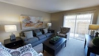 This single-floor space offers a comfortable and thoughtfully designed living environment. The living room serves as the focal point of the home, featuring plush carpeted flooring and neutral-toned walls that create a warm, inviting atmosphere. A large sliding glass door with vertical blinds floods the room with natural light while providing direct access to an outdoor area, enhancing the sense of openness and connectivity to the exterior.

The living room is anchored by a generously sized gray sofa complemented by coordinating accent chairs with decorative patterned upholstery, arranged around a dark-finished rectangular coffee table. A bold, colorful abstract painting adorns the main wall, adding a vibrant artistic touch to the otherwise neutral palette. Subtle, well-placed lighting, including a classic table lamp, contributes to the room's relaxed ambiance.

The overall design aesthetic is modern yet approachable, blending contemporary furnishings with warm, earthy tones. The space is well-proportioned, offering a comfortable flow that is both functional and visually appealing — making it an ideal environment for relaxing or entertaining.