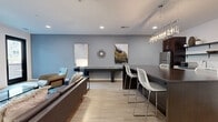 This well-appointed amenity space offers a sophisticated blend of comfort and functionality within a thoughtfully designed single-floor layout. Light wood-tone flooring runs throughout, complementing the cool blue-gray and crisp white wall palette that lends the space a calm, modern aesthetic.

The main lounge area is anchored by plush leather seating, accent chairs, and a glass-topped coffee table, all bathed in natural light from large black-framed windows. A wall-mounted flat-screen TV and lush tropical plant add character and warmth to the gathering area.

Adjacent to the lounge, a sleek kitchenette is outfitted with rich dark wood cabinetry, open metal shelving, a stainless steel refrigerator, microwave, and sink — all centered around a substantial dark-topped island with bar-height seating. A cascading crystal-style pendant chandelier adds an elegant focal point overhead.

A billiards table positioned along the back wall provides a recreational anchor to the space, while curated wall art and decorative accents enhance the contemporary ambiance throughout.

A separate, more intimate meeting or dining room features a striking marble-topped table, modern white chairs, and bold artwork, offering a quieter setting for focused gatherings. Recessed lighting throughout ensures consistent, well-distributed illumination across all zones.