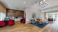 This contemporary community space boasts a spacious, open-concept layout with soaring ceilings and abundant natural light. The main area features a comfortable lounge with a tufted gray sofa, vibrant artwork, and a blue area rug. Adjacent is a dining area with a round table and red metal chairs, ideal for gatherings. A sleek kitchenette with white cabinetry and stainless steel appliances offers convenience. The space is enhanced by warm wood flooring throughout, creating a cohesive and inviting atmosphere. Large windows and glass doors provide views of an outdoor pool area, seamlessly connecting indoor and outdoor spaces.