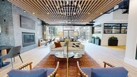 Located at 2200 Market St in Denver's vibrant Curtis Park neighborhood, this amenity level combines a game room, WiFi lounge, and outdoor courtyard into a cohesive, well-appointed communal space. The interior features an industrial-modern aesthetic, with exposed ductwork, track lighting, and light hardwood flooring throughout. The game room is anchored by two full-size billiards tables beneath globe pendant lights, set against a bold neon "Premier Tire Terminal" wall sign that nods to the site's history. Slatted wood dividers define zones without closing off the open floor plan. A built-in kitchenette with subway tile backsplash and a large-format refrigerator supports entertaining, while a richly stained bar-height island provides casual seating. The WiFi lounge offers semi-private booth seating with tan leather banquettes framed by sleek black steel-and-glass partitions, ideal for focused work or relaxed conversation. The adjacent outdoor courtyard extends the living space with generous white sectional sofas flanking a linear fire pit table, a water feature wall, lush vertical greenery, and café-style seating — all open to the Colorado sky above.