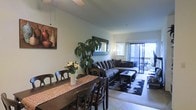 This single-floor space offers a thoughtfully arranged open-plan layout that seamlessly connects the dining and living areas, creating a cohesive and comfortable environment. The dining area accommodates a generously sized dark wood table with seating for six, accented by a decorative table runner and a fresh floral centerpiece. A large indoor plant adds a vibrant, natural touch to the space, enhancing the warm and inviting ambiance.

The living area is anchored by a plush sectional sofa arranged around a rectangular coffee table, set atop a bold patterned area rug that adds visual interest to the tiled flooring. A decorative mirror and framed artwork on the walls contribute to the curated aesthetic of the space.

One of the standout features is the sliding glass door that opens directly onto a private balcony, flooding the interior with natural light and offering a seamless indoor-outdoor connection. The light-toned walls and neutral palette throughout keep the space feeling open and airy. The overall ambiance is relaxed yet polished, making it well-suited for both everyday living and casual entertaining.