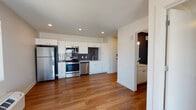 This single-floor unit offers a clean, modern aesthetic throughout, combining functional design with contemporary finishes. The open-plan kitchen and living area serves as the heart of the space, anchored by rich hardwood-style flooring that runs throughout. The kitchen is well-appointed with a full suite of stainless steel appliances, including a refrigerator, electric range, over-the-range microwave, and dishwasher. White flat-panel cabinetry provides ample storage, while a slate-toned tile backsplash adds a stylish contrast. A stainless steel sink with a modern pull-down faucet and light-colored countertops complete the workspace. Recessed lighting illuminates the area efficiently, and a window brings in natural light with views of the surrounding urban environment. The living area is open and airy, with a wall-mounted climate control unit ensuring year-round comfort. The bedroom is a quiet, comfortable retreat featuring plush carpeting, neutral walls, and a large window that frames city views and fills the room with natural light. The bathroom is thoughtfully designed with wood-look flooring, a white vanity with a single sink, a full tub-and-shower combination, and a bold dark accent wall that adds a modern touch to the otherwise bright and clean space.
