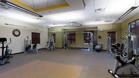 This fitness center is a single-floor facility designed to accommodate a comprehensive workout experience. The space is defined by its open layout, offering generous room for both strength training and open-floor exercises. The workout area features multiple cable and resistance machines arranged throughout the room, providing a variety of strength training options for users of all fitness levels. A stability ball and additional equipment round out the training options available.

The interior is finished with a combination of durable carpet and rubberized flooring, providing appropriate surfaces for both machine-based and floor exercises. The walls are painted in a warm olive green tone, accented by bold yellow architectural details near the ceiling that add a vibrant, energetic character to the space. A drop ceiling with recessed panel tiles houses fluorescent lighting fixtures that ensure the room is consistently and evenly illuminated throughout.

Several windows fitted with dark wood blinds allow for natural light while maintaining privacy. Ceiling fans are installed to promote air circulation, contributing to a comfortable environment during workouts. The overall ambiance is functional and purposeful, offering a clean, organized, and well-maintained environment suited for daily fitness activity.