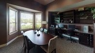 This single-floor space presents a refined and functional environment designed for professional use. The interior is defined by deep, warm brown walls that create a sophisticated atmosphere throughout. The main area seamlessly integrates a conference zone and a dedicated workstation area, making it a versatile setting for both meetings and daily business operations.

The conference area centers around a rich, dark cherry oval table surrounded by leather and upholstered chairs. A striking bay window arrangement floods the space with natural light while offering views of the surrounding greenery, complemented by adjustable blinds for privacy. A tall, decorative floor lamp adds a warm ambient glow.

The built-in workstation along one wall features a long desk surface accommodating multiple computer monitors, paired with dark cabinetry above and below offering ample storage. Open shelving units display decorative greenery and accessories, contributing to the polished aesthetic. A dark wood glass-front cabinet adds additional storage and visual interest.

Patterned carpeting runs throughout, grounding the space, while framed artwork adorns the walls. The overall design balances practicality with an upscale, welcoming ambiance suited to a professional setting.