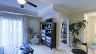 This single-floor residence offers a thoughtfully designed living space that balances comfort and style. The main living area immediately draws the eye upward to its distinctive tray ceiling, accented by crisp crown molding and complemented by a ceiling fan that adds both function and charm. Neutral, warm wall tones create a welcoming ambiance throughout, while plush light-colored carpeting underfoot adds a sense of warmth and comfort.

The living room is generously proportioned, providing ample space for both relaxation and entertainment. A dark-finished armoire serves as a striking focal point, offering display shelving and storage, and pairing elegantly with the room's overall color palette. A sleek glass-topped coffee table and a light blue accent chair add modern, curated touches to the space.

An open passageway connects the living area to an adjoining room — likely a home office or den — where built-in shelving lines the walls, offering organized storage and display space. Large windows dressed with flowing white curtains invite natural light to fill the interior, enhancing the bright and airy feel of the home. Decorative greenery and tall dried botanicals are thoughtfully placed, adding organic texture and visual interest. The overall layout is open and fluid, making this a versatile and inviting single-level home.
