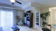 This single-floor residence offers a thoughtfully designed living space that balances comfort and style. The main living area immediately draws the eye upward to its distinctive tray ceiling, accented by crisp crown molding and complemented by a ceiling fan that adds both function and charm. Neutral, warm wall tones create a welcoming ambiance throughout, while plush light-colored carpeting underfoot adds a sense of warmth and comfort.

The living room is generously proportioned, providing ample space for both relaxation and entertainment. A dark-finished armoire serves as a striking focal point, offering display shelving and storage, and pairing elegantly with the room's overall color palette. A sleek glass-topped coffee table and a light blue accent chair add modern, curated touches to the space.

An open passageway connects the living area to an adjoining room — likely a home office or den — where built-in shelving lines the walls, offering organized storage and display space. Large windows dressed with flowing white curtains invite natural light to fill the interior, enhancing the bright and airy feel of the home. Decorative greenery and tall dried botanicals are thoughtfully placed, adding organic texture and visual interest. The overall layout is open and fluid, making this a versatile and inviting single-level home.