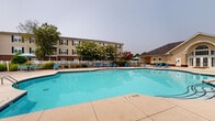 Located at 1025 Kensington Dr in Jacksonville, North Carolina, this outdoor community amenity space offers a well-appointed recreational area designed for residents to relax and socialize. At the heart of the space is a generously sized swimming pool with a curved, freeform shape, featuring clearly marked depth indicators and equipped with stainless steel entry rails for easy access. The surrounding concrete sun deck provides ample room for lounging, and is furnished with multiple blue lounge chairs and umbrellas positioned throughout, offering both sun and shade options. A white perimeter fence encloses the pool area, creating a defined and secure environment. Adjacent to the pool is a clubhouse building with a distinctive gabled roofline and warm yellow siding, providing a dedicated facility for community use. Mature trees and ornamental plantings, including flowering varieties, add natural beauty and shade to the landscape. A playground area is visible beyond the pool enclosure, offering recreational space for younger residents. The multi-story residential buildings, featuring classic brick and neutral-toned exteriors with black-shuttered windows, frame the backdrop of the amenity area, contributing to a cohesive and inviting community atmosphere.